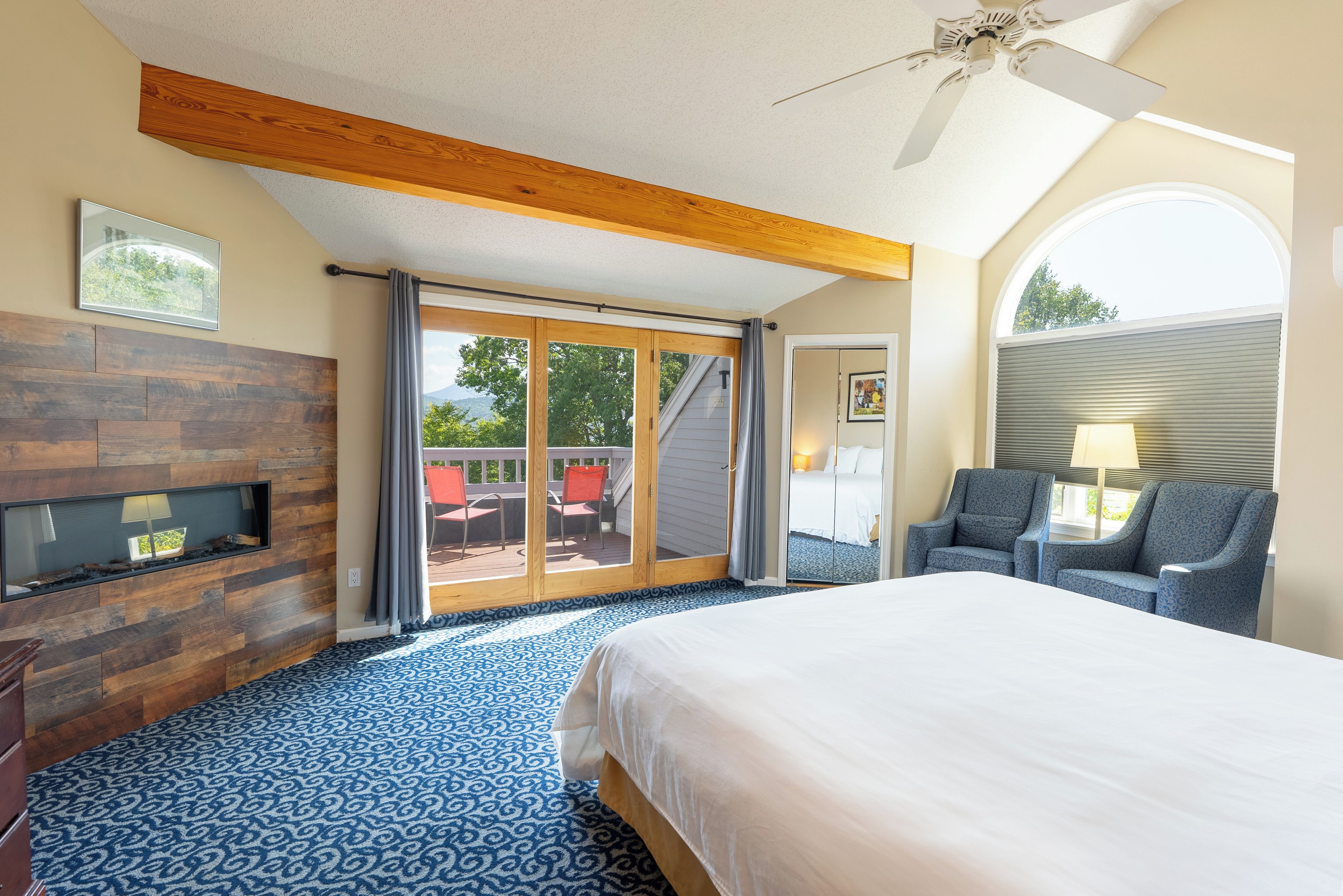 A hotel room with a large bed, a TV mounted on a wooden panel wall, a ceiling fan, and a view of the outdoors through a sliding glass door.