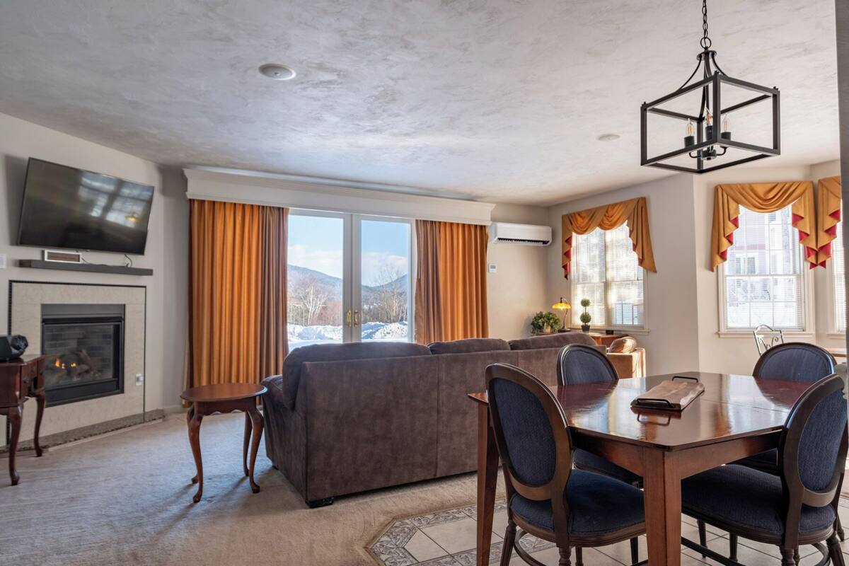 A living room with a fireplace, a television, a sofa, a dining table with chairs, and large windows with curtains.