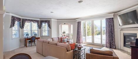 A spacious living room with a large sofa, a coffee table, and a fireplace. There are large windows with curtains, a dining area, and a view of the outdoors.