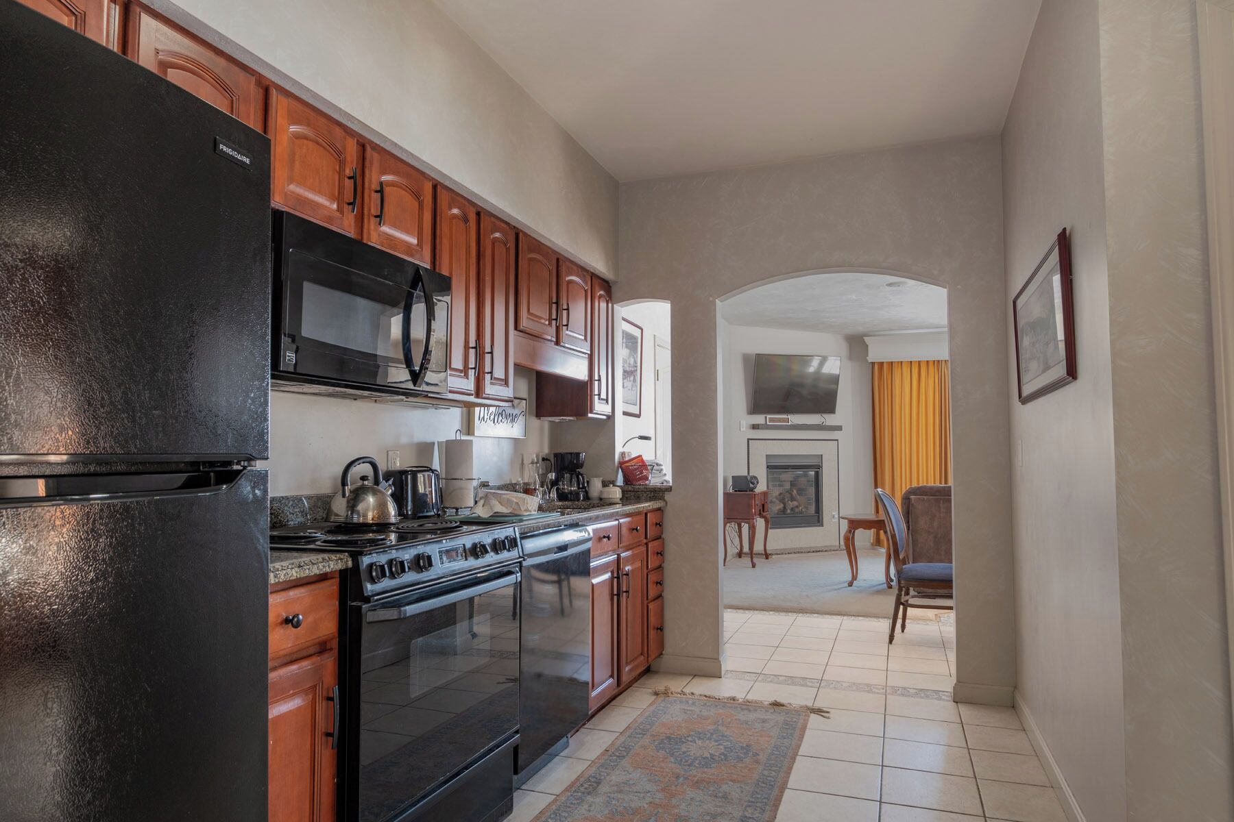 A kitchen with wooden cabinets, a black refrigerator, a stove, and a microwave. There is a rug on the floor and a dining area visible in the background.