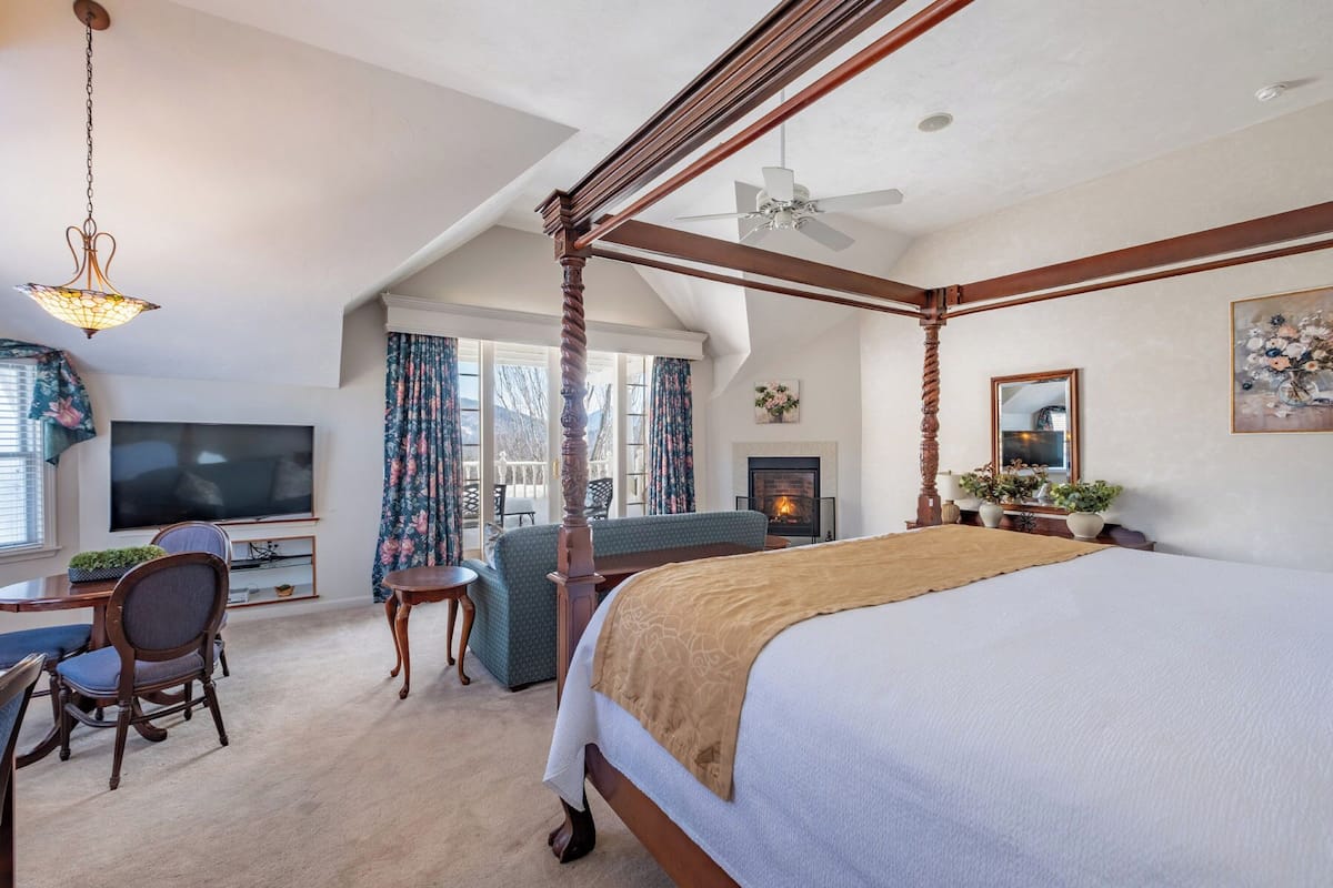 A spacious bedroom with a four-poster bed, a television, a desk with a chair, and a fireplace.