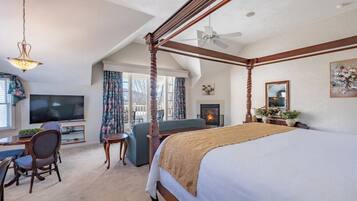 A spacious bedroom with a four-poster bed, a television, a desk with a chair, and a fireplace.