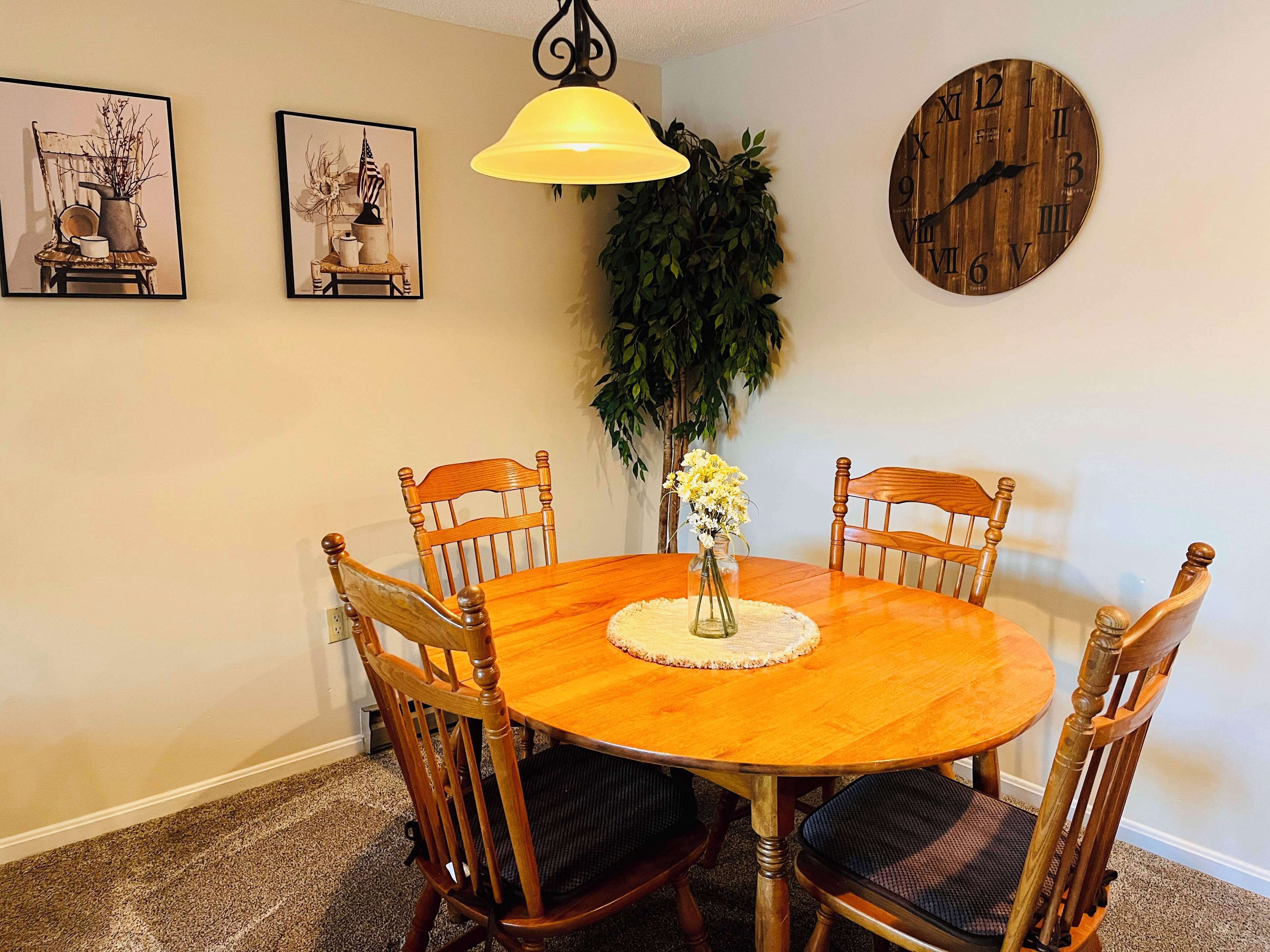 A dining room with a wooden table, four chairs, a yellow pendant light, and two wall-mounted clocks.