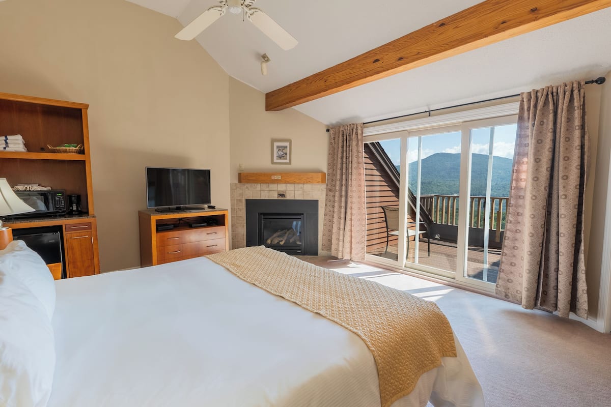 A hotel room with a bed, a television, a fireplace, a wooden shelf, and a balcony with a view of the mountains.