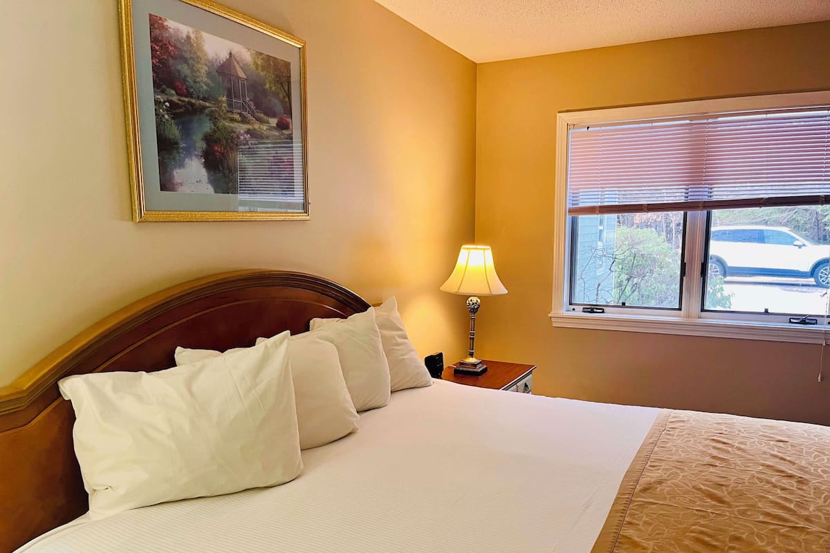 A neatly made bed with white linens and pillows, a bedside lamp, a framed picture on the wall, and a window with blinds.