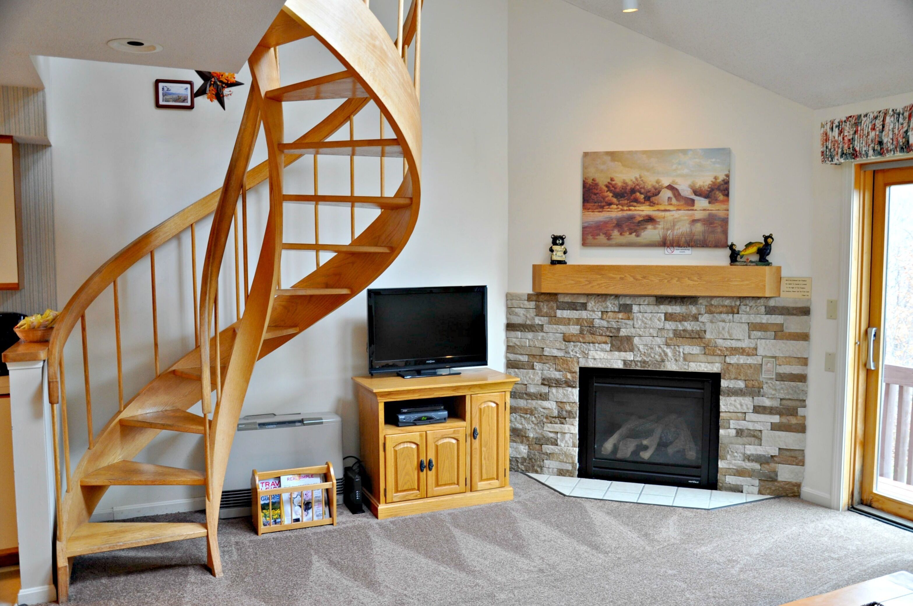 Ein Wohnzimmer mit einer Wendeltreppe aus Holz, einem Flachbildfernseher, einem Kamin aus Stein und einem Gemälde über dem Kaminvorsatz.