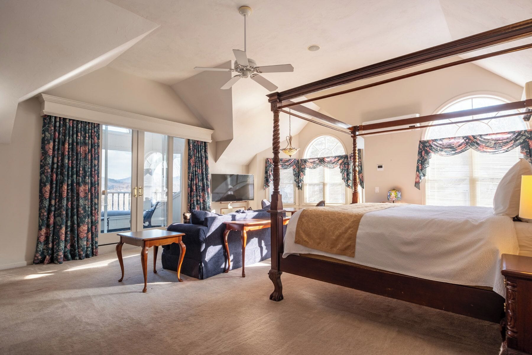 A four-poster bed with a canopy, a desk with a chair, a television, and a view of the outdoors through large windows.