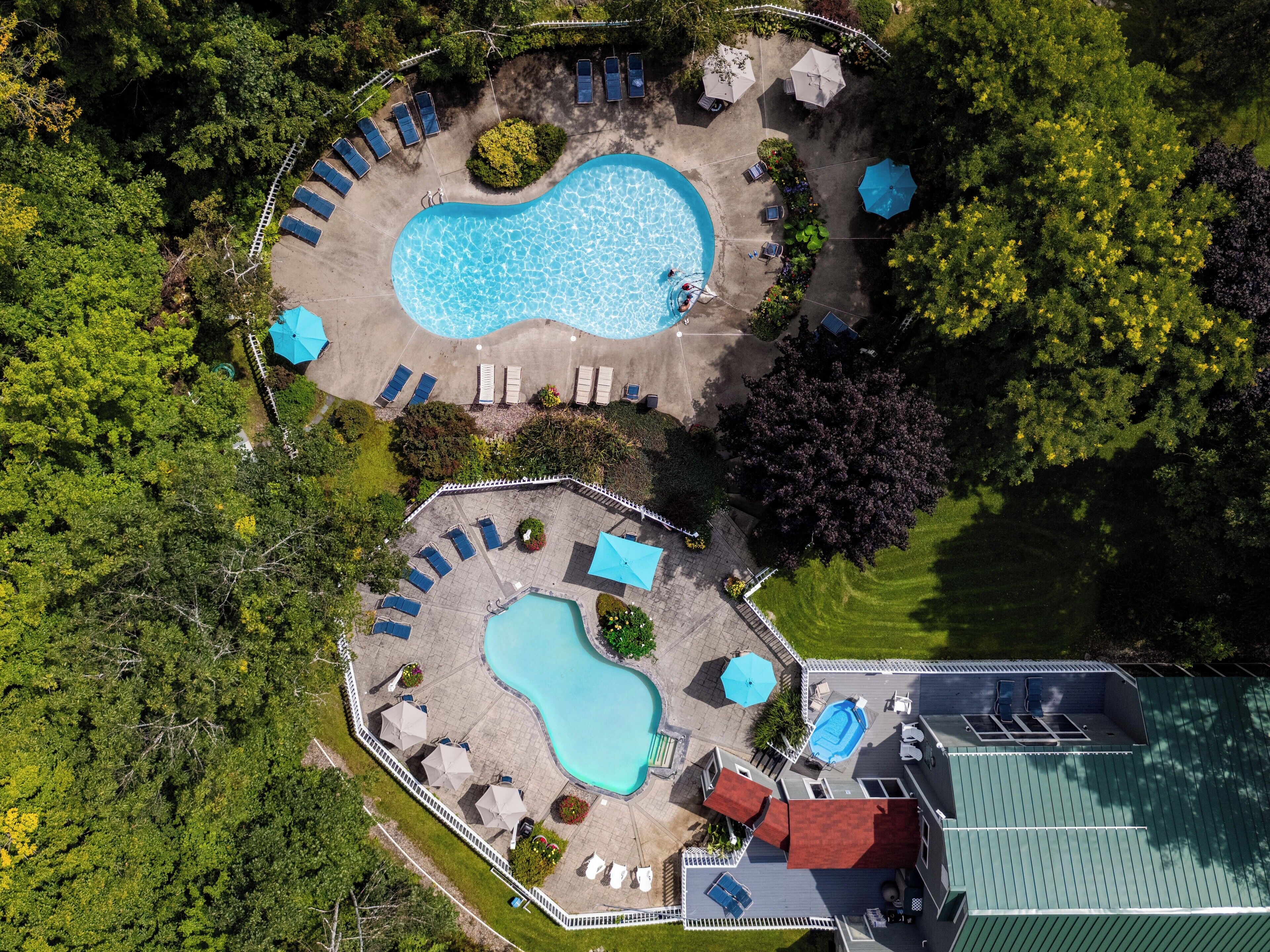An aerial view of a resort with two swimming pools, sun loungers, umbrellas, and a fenced area with a building.
