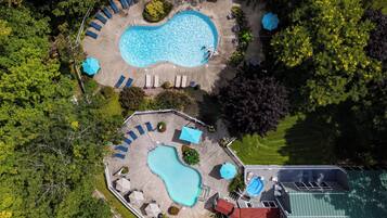 An aerial view of a resort with two swimming pools, sun loungers, umbrellas, and a fenced area with a building.