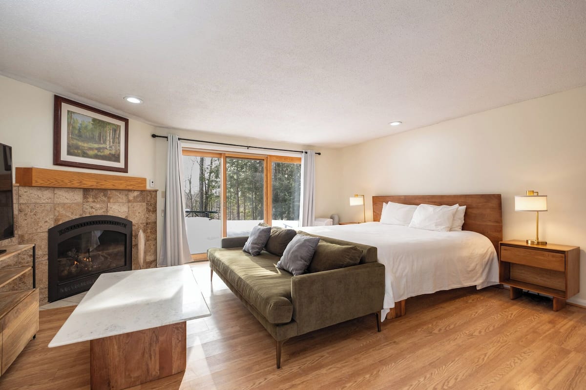 A bedroom with a bed, a sofa, a nightstand, a lamp, a fireplace, and a large window.