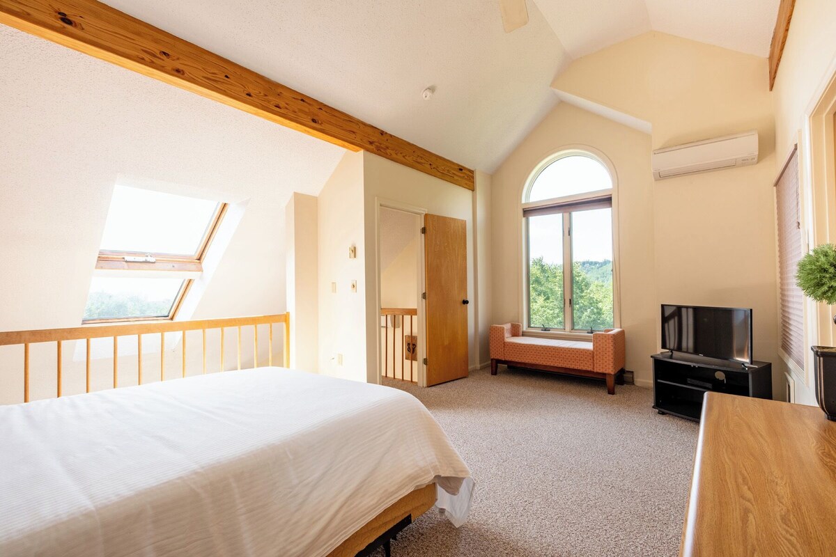 A bedroom with a bed, a bench, a TV, and a window with a view.