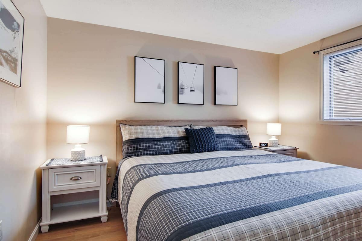 A bedroom with a bed, two bedside tables, a nightstand, a window with blinds, and three framed pictures on the wall.