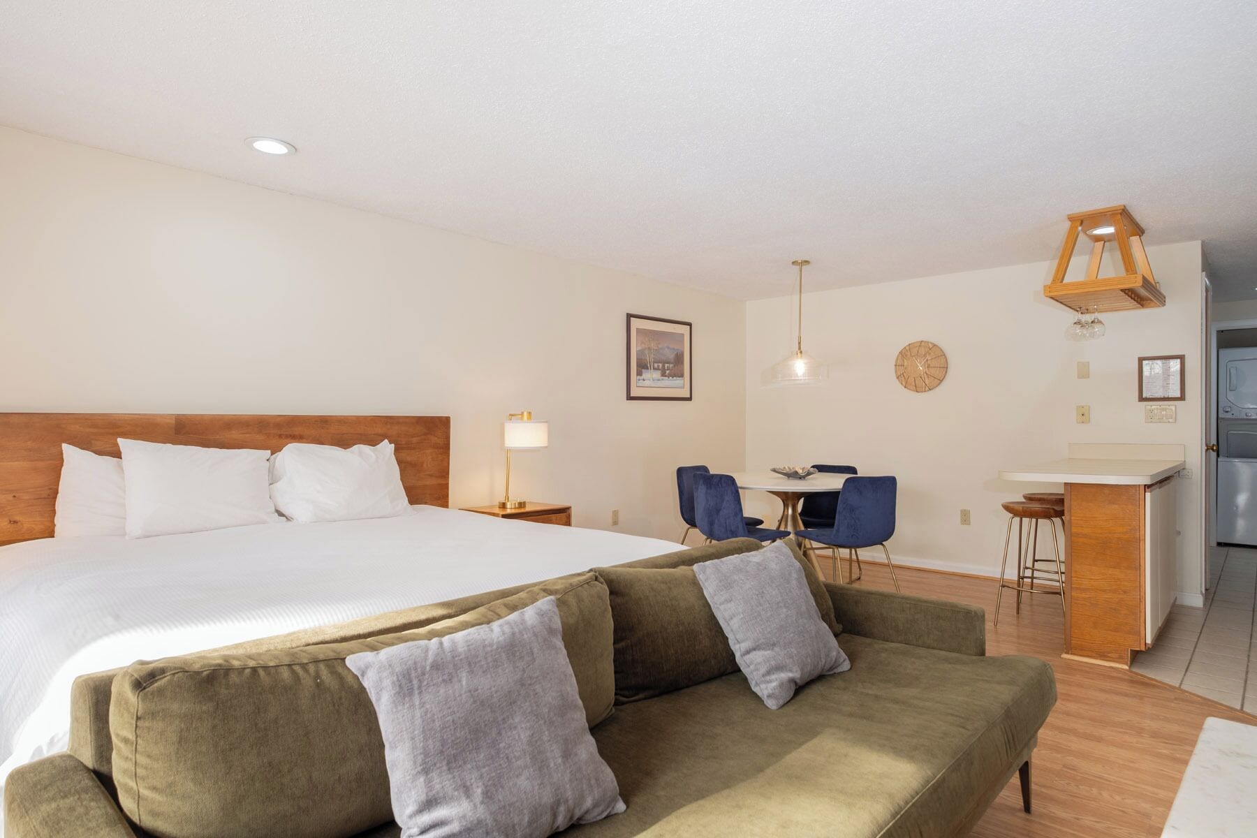 A hotel room with a large bed, a sofa, a dining table with chairs, a kitchen area, and a wall-mounted clock.