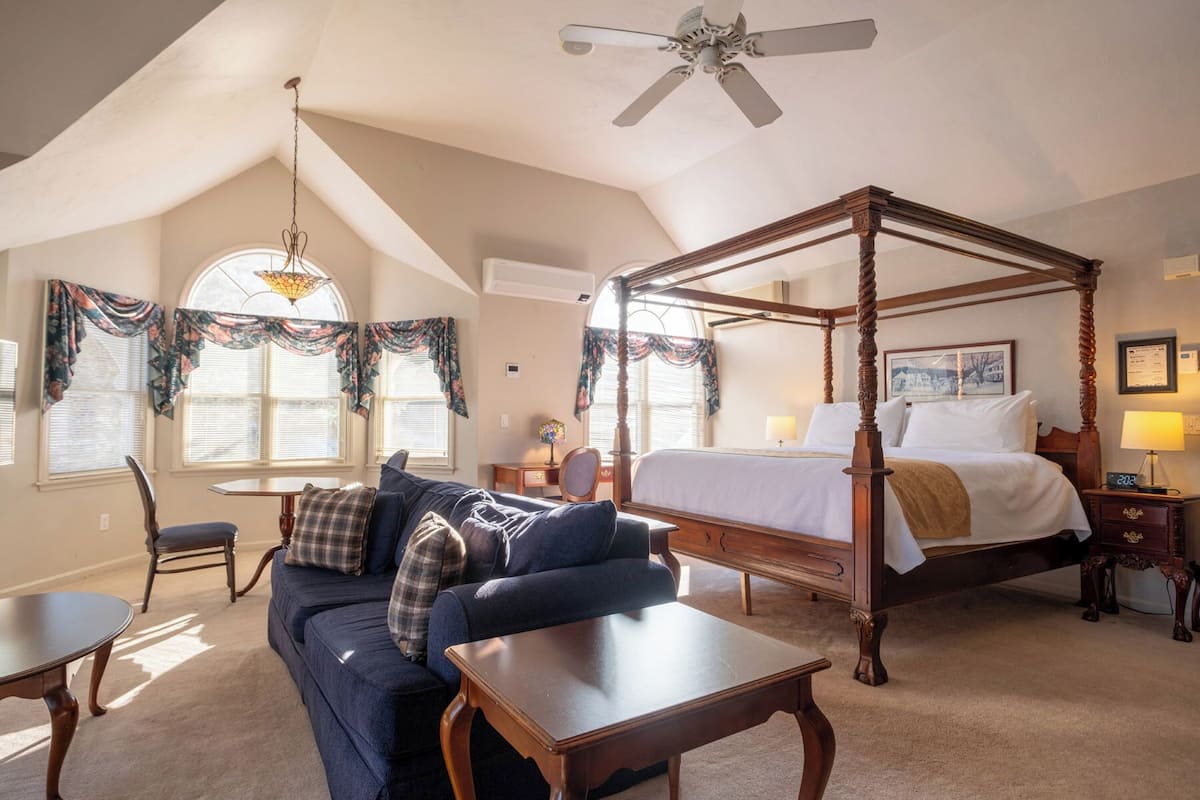 A bedroom with a four-poster bed, a sitting area with a sofa and armchair, a chandelier, and a ceiling fan.