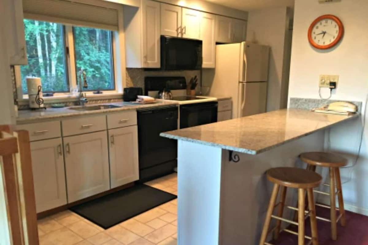 A kitchen with white cabinets, a black microwave, a refrigerator, and a window with a view of trees. There are two wooden stools and a black doormat.
