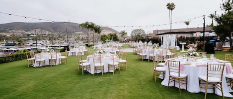 An outdoor wedding venue with multiple round tables set up on a grassy lawn, surrounded by white chairs and draped with white tablecloths.