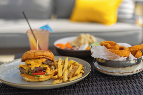 Un plato con una hamburguesa y papas fritas, un bol de aros de cebolla y una bebida con pajita sobre una mesa.