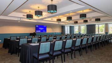 A conference room with rows of tables and chairs, a large screen displaying a presentation, and a view of the outdoors through large windows.