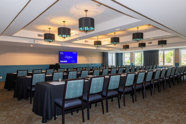 Un salón de conferencias con filas de mesas y sillas, una pantalla grande mostrando una presentación, y vistas al exterior a través de amplias ventanas.