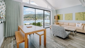 A spacious living area with a wooden table, a sofa, and a chair, overlooking a scenic view of a body of water and hills.