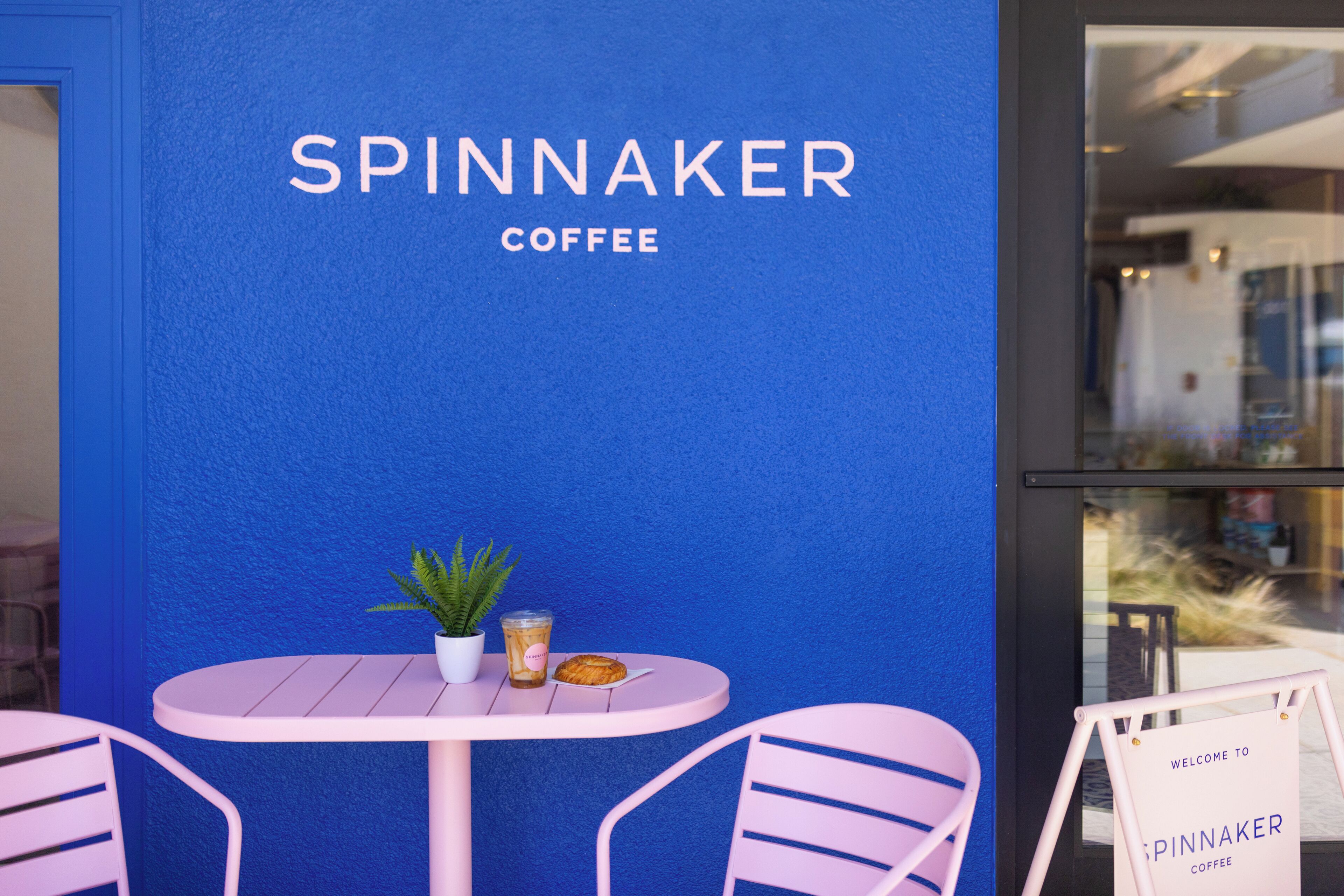 Pared azul con el texto "SPINNAKER COFFEE" y una pequeña mesa con una planta en maceta, un vaso y un pastel.