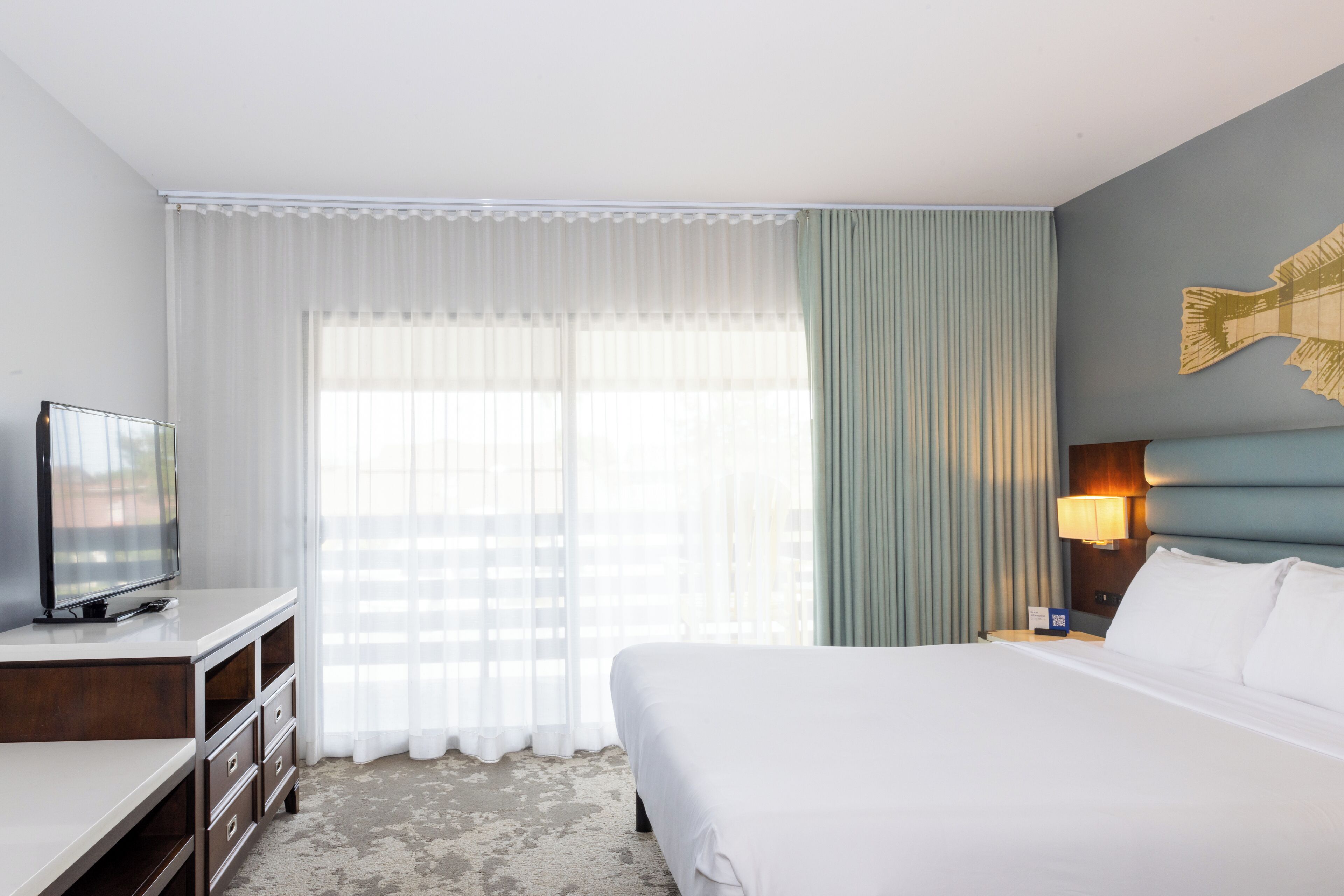 A hotel room with a bed, a TV on a desk, a lamp, and a window with curtains.
