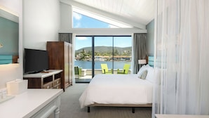 A hotel room with a large bed, a TV, a desk with a lamp, and a view of a lake and mountains.