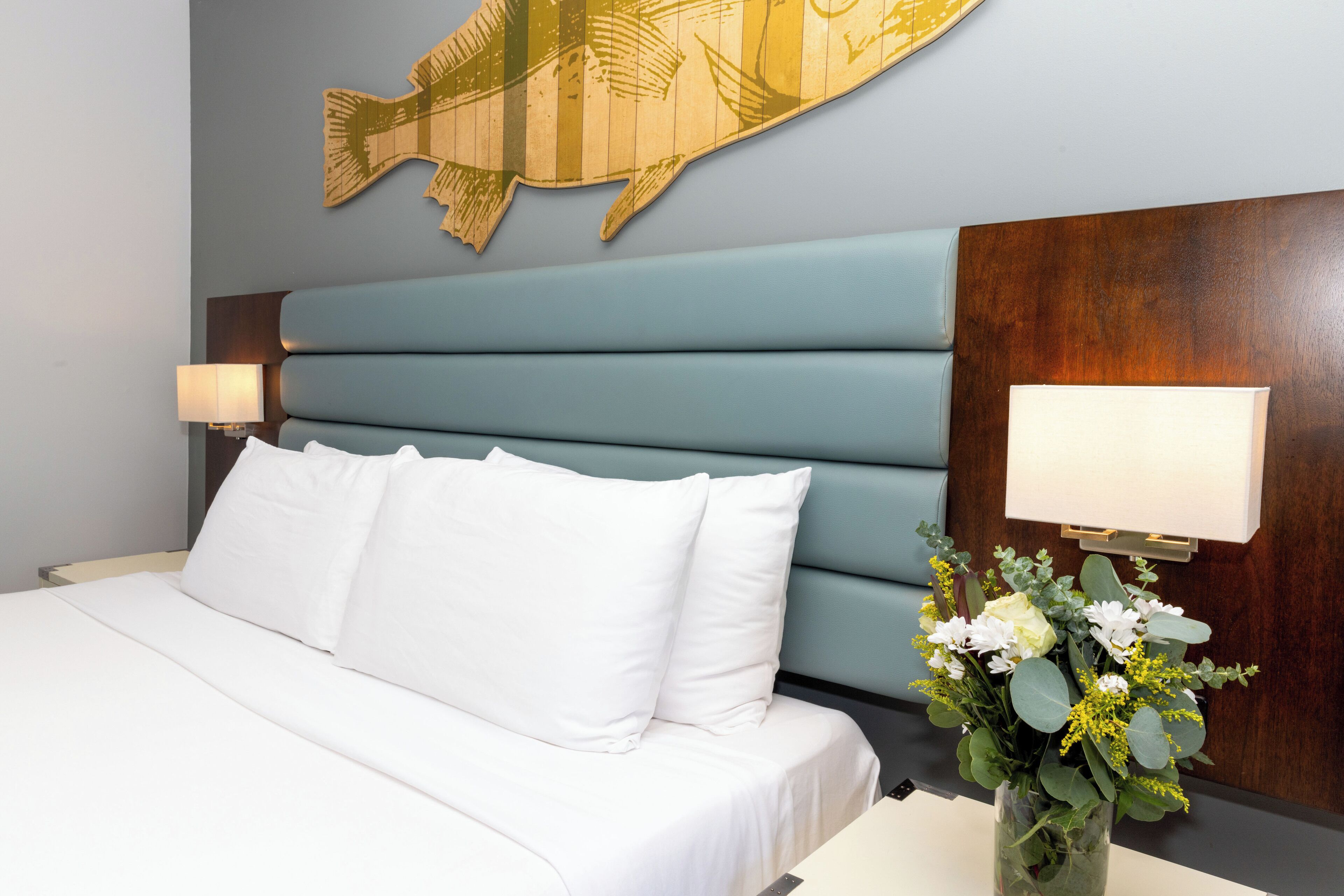Een hotelkamer met een bed, witte lakens, een vaas met bloemen en een kunstwerk aan de muur.