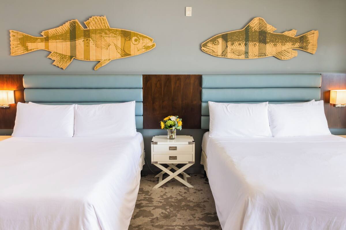 Een hotelkamer met twee bedden, een houten vis-wanddecoratie, een nachtkastje met een bloevas en een houten hoofdbord.