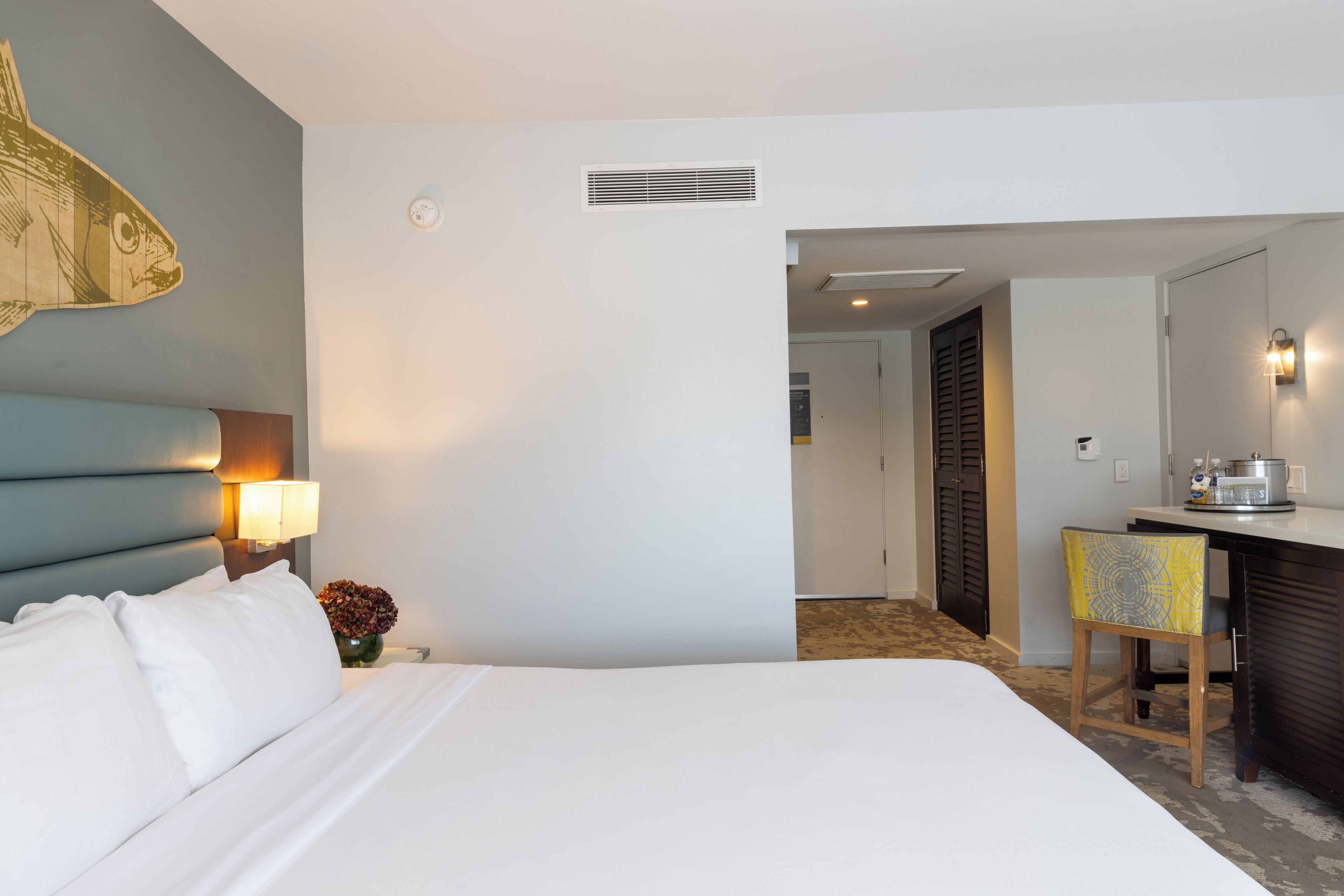 A hotel room with a large bed, a desk with a chair, a wall-mounted lamp, and a decorative fish wall art piece.
