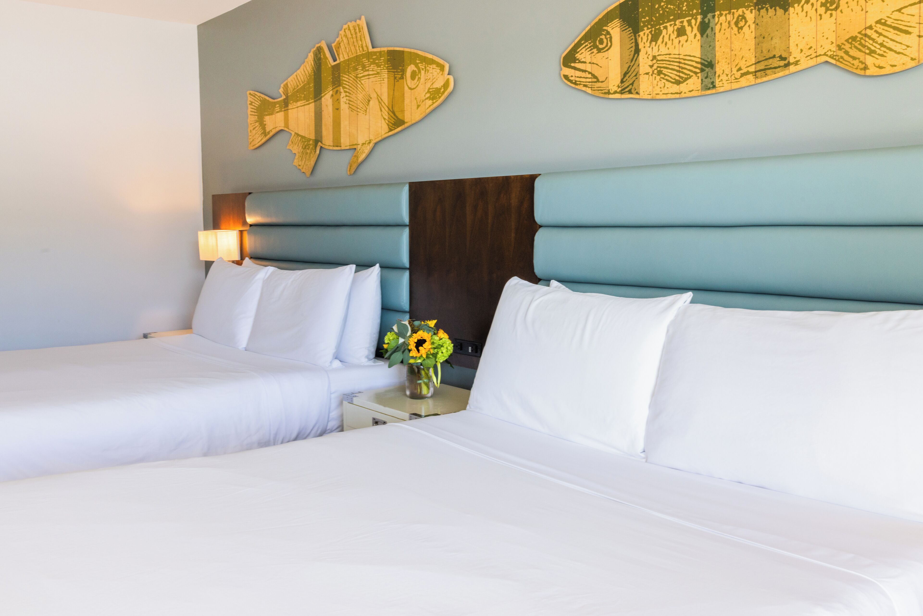 Habitación de hotel con dos camas, una mesita de noche con un jarrón de flores y cuadros de peces.