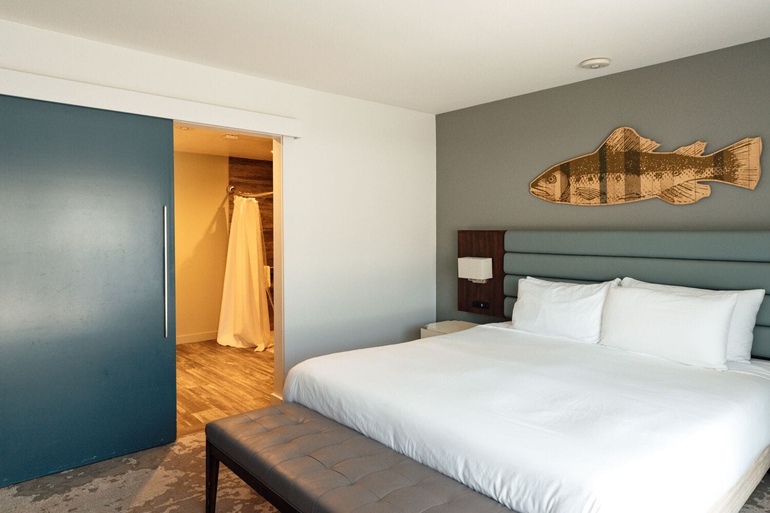 A hotel room with a bed, a bench, a wall-mounted fish artwork, and a door leading to a bathroom.