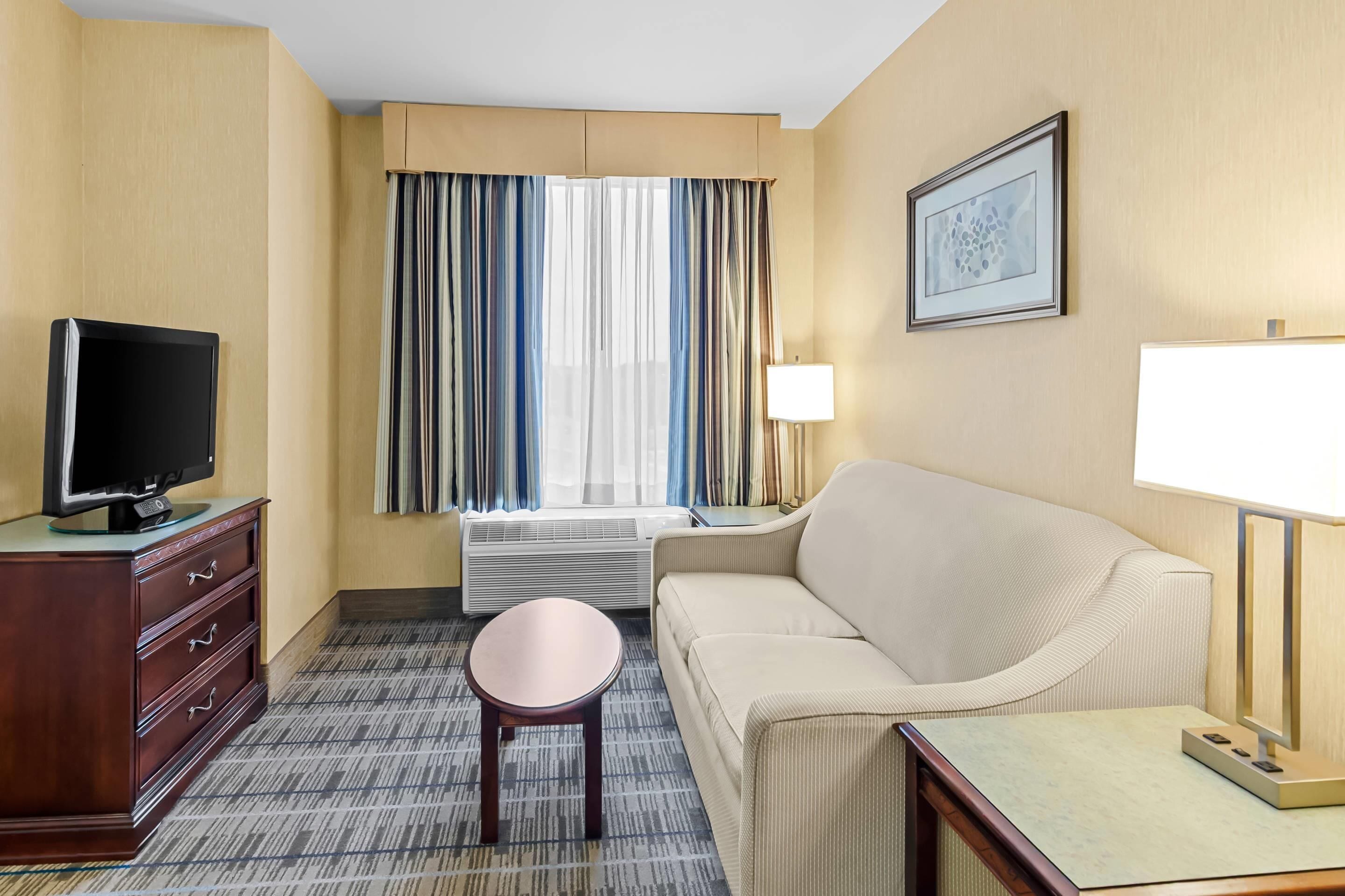 A hotel room with a flat-screen TV on a wooden dresser, a beige sofa, a small round table, a standing lamp, and a window with striped curtains.