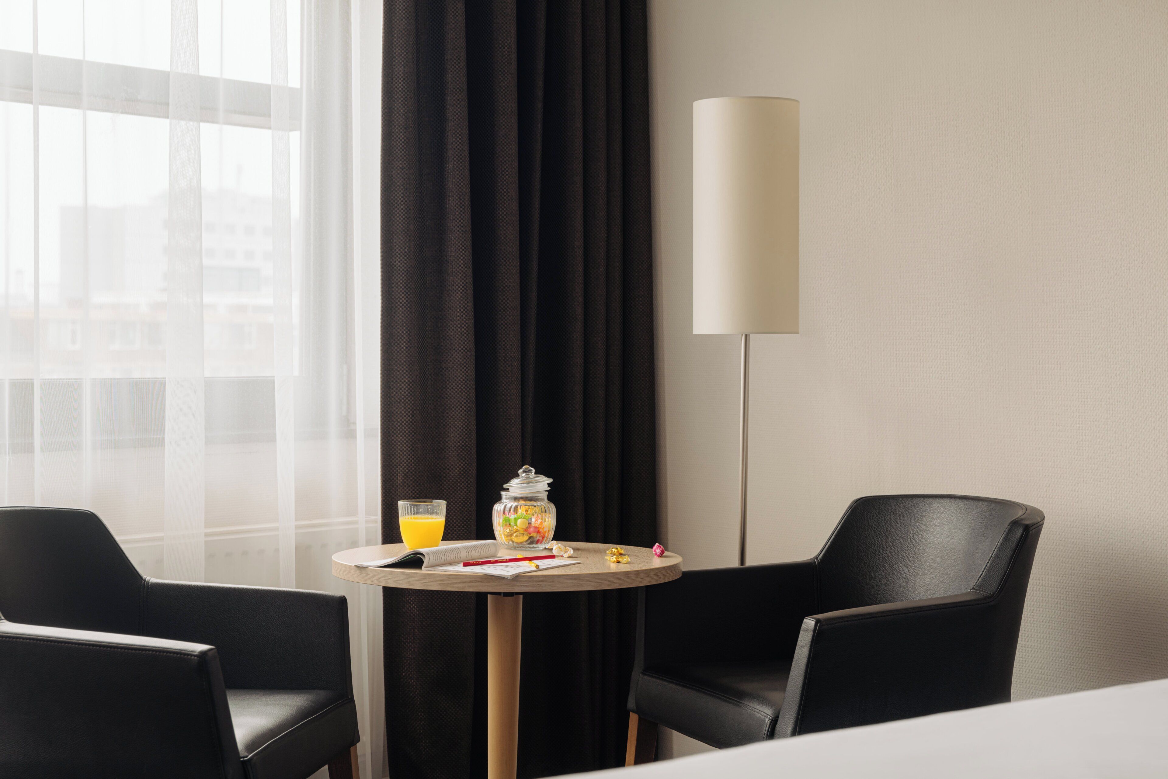Habitación de hotel con una mesa redonda, dos sillas negras, una lámpara, un vaso de jugo de naranja y un frasco de aperitivos.