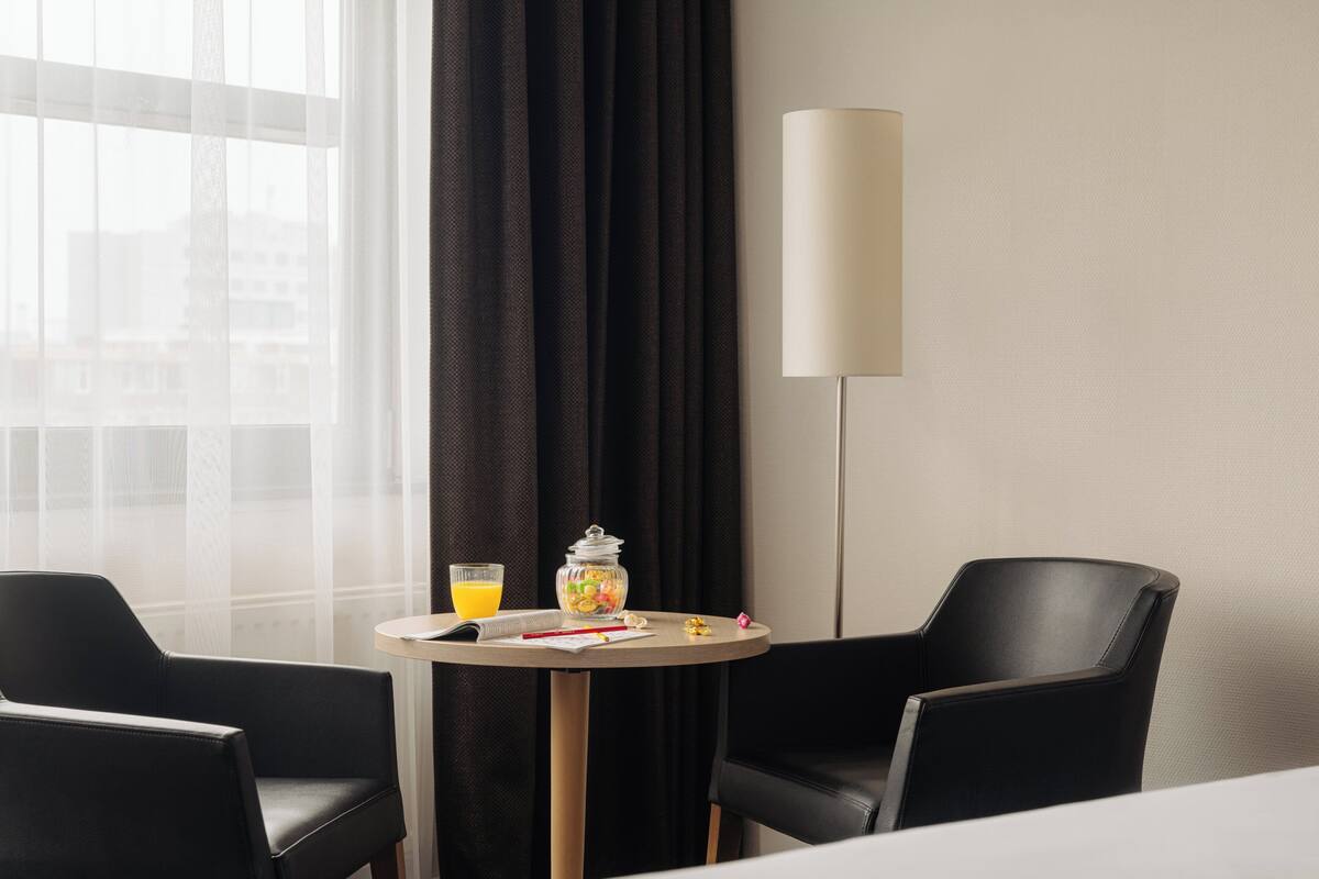 A hotel room with a round table, two black chairs, a lamp, a glass of orange juice, and a jar of snacks.