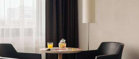 Habitación de hotel con una mesa redonda, dos sillas negras, una lámpara, un vaso de jugo de naranja y un frasco de aperitivos.