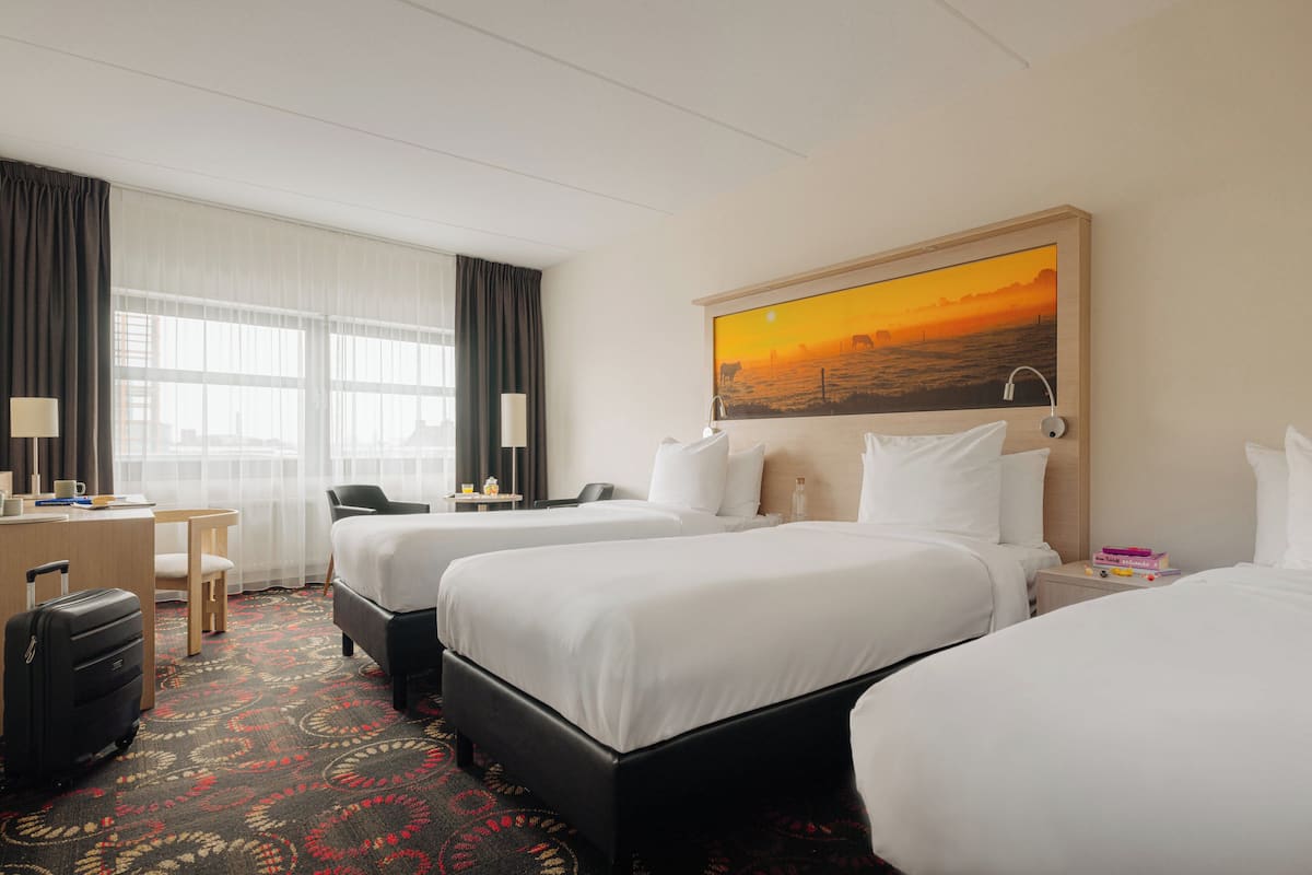 A hotel room with two beds, a desk, a chair, a suitcase, and a painting of a sunset.