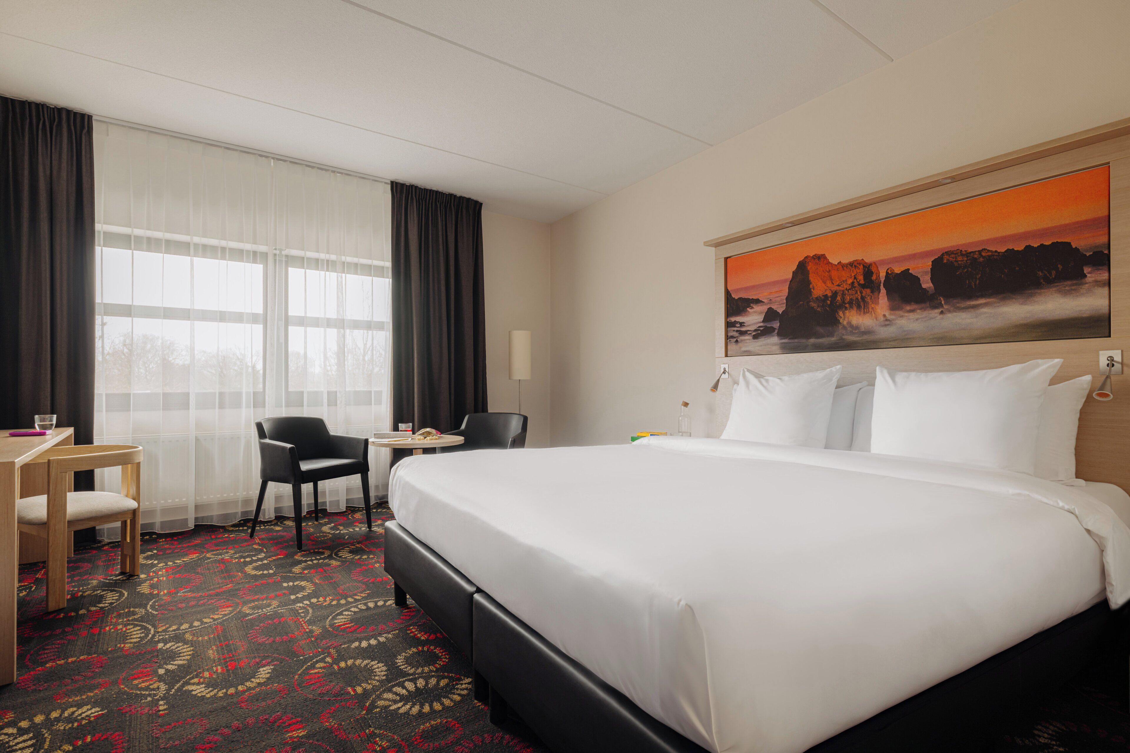 Une chambre d’hôtel avec un grand lit, un bureau avec une chaise, une petite table et une grande fenêtre avec des rideaux.