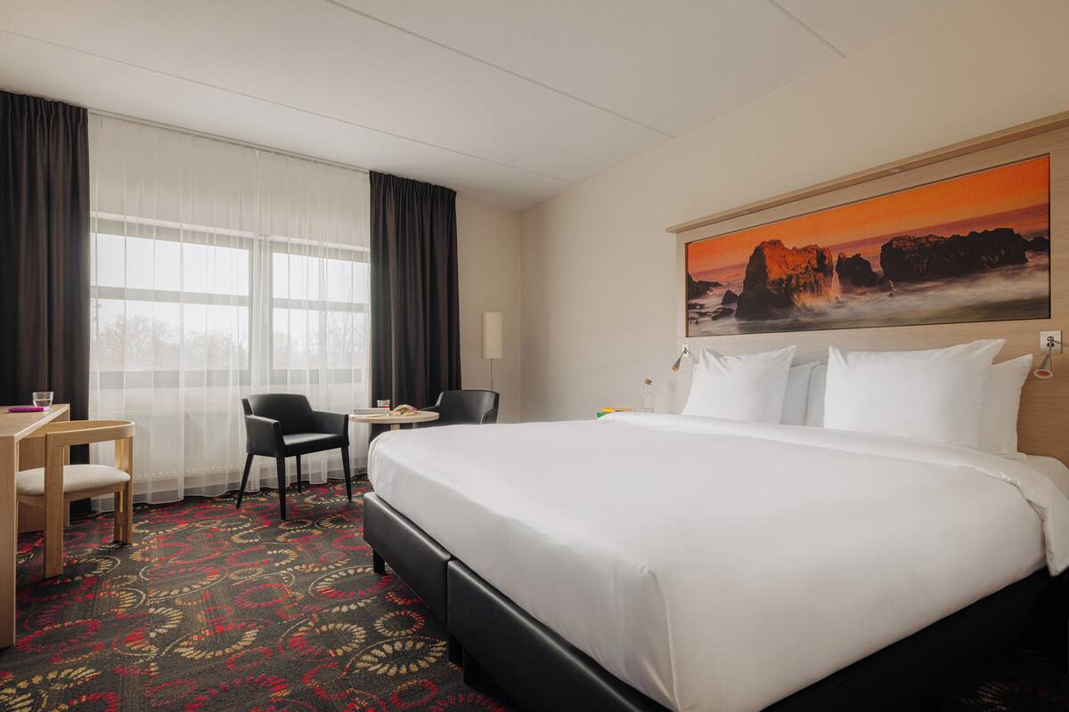 A hotel room with a large bed, a desk with a chair, a small table, and a large window with curtains.