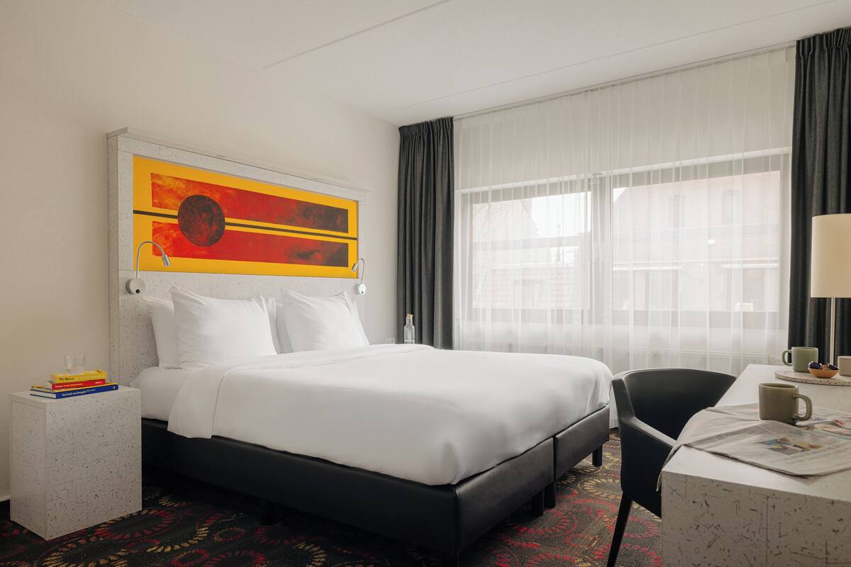 A hotel room with a large bed, a side table with books, a desk with a chair, and a window with sheer curtains.