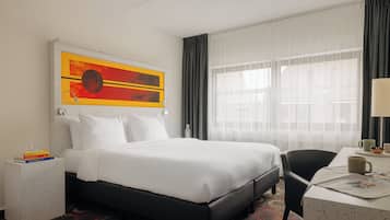 Una habitación de hotel con una cama grande, una mesita de noche con libros, un escritorio con una silla y una ventana con cortinas translúcidas.