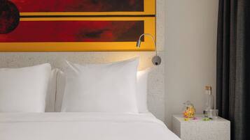 Habitación de hotel con cama, almohadas blancas, mesita de noche con lámpara, un adorno en la pared y una pequeña mesa con un tarro de caramelos.