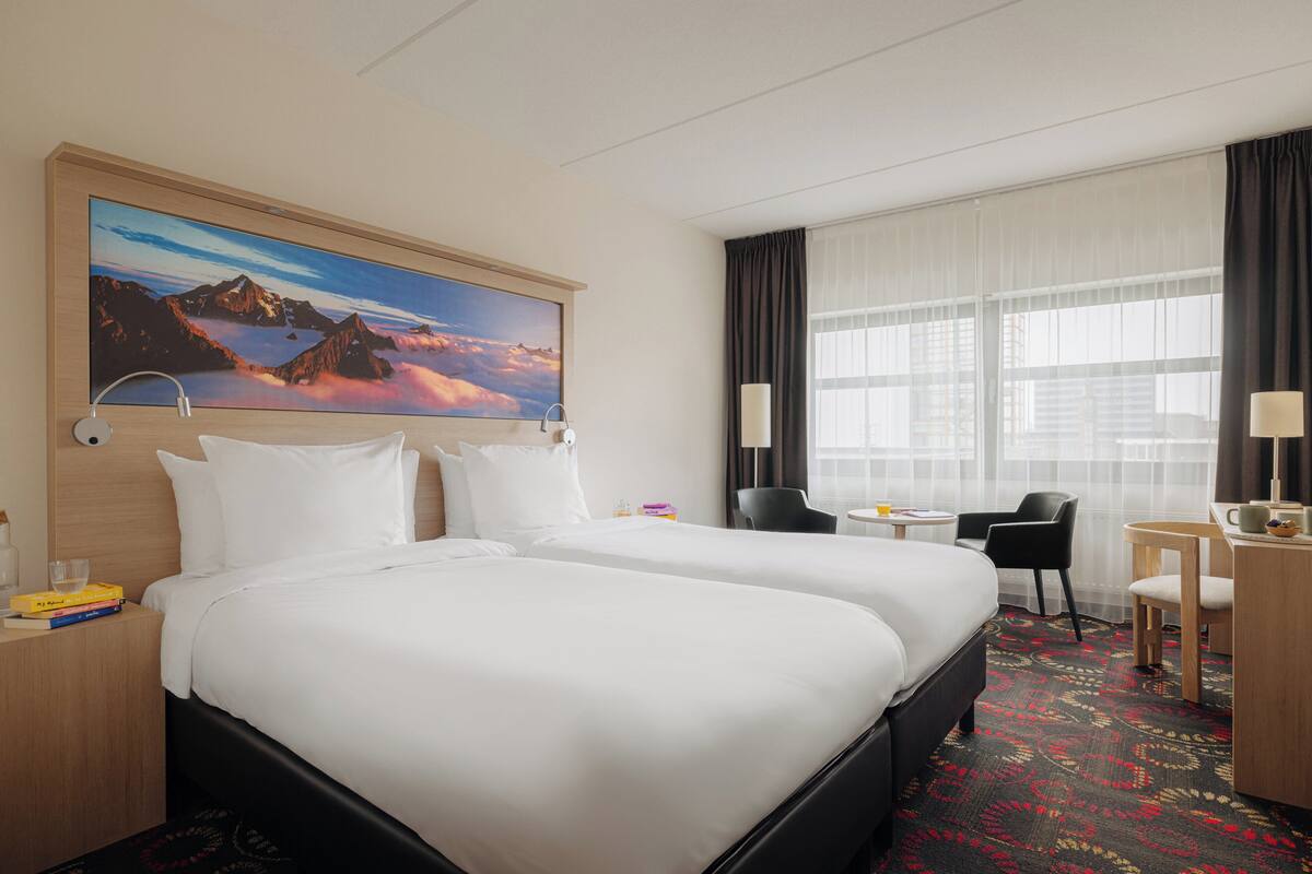 A hotel room with two beds, a desk, a chair, a large window with curtains, and a painting on the wall.
