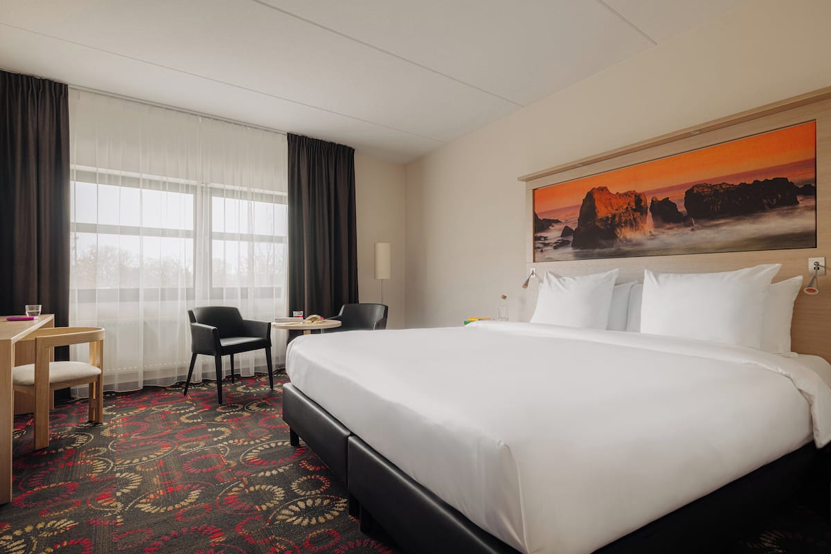 A hotel room with a large bed, a desk with a chair, a small table, and a large window with curtains.