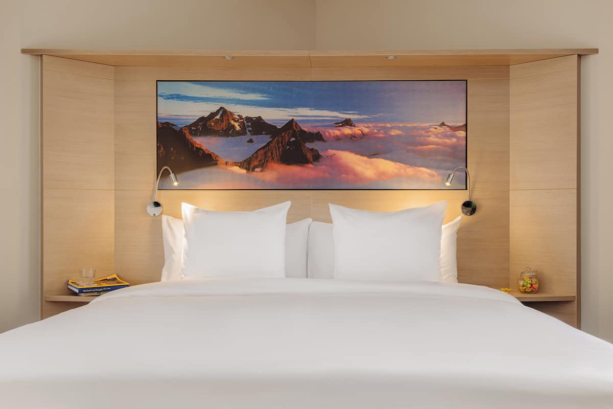 A hotel room with a large bed, white bedding, a mounted artwork of mountains and clouds, and a bedside table with a glass and a book.