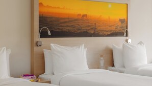 A hotel room with a large painting of a sunset over a field, a wooden headboard, white bedding, and a bedside table with a bottle and a book. - Leonardo Hotel Hengelo City Center (Hengelo)