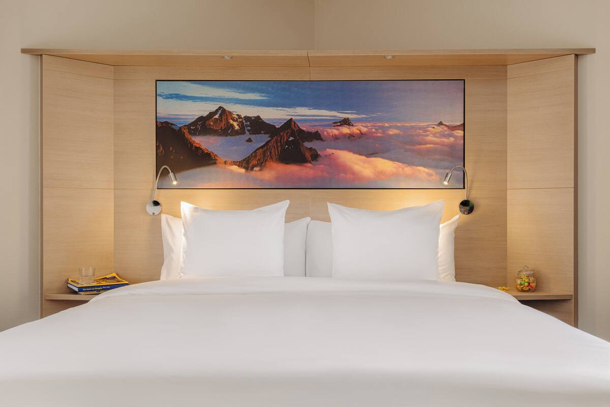 A hotel room with a large bed, white bedding, a mounted artwork of mountains and clouds, and a bedside table with a glass and a book.