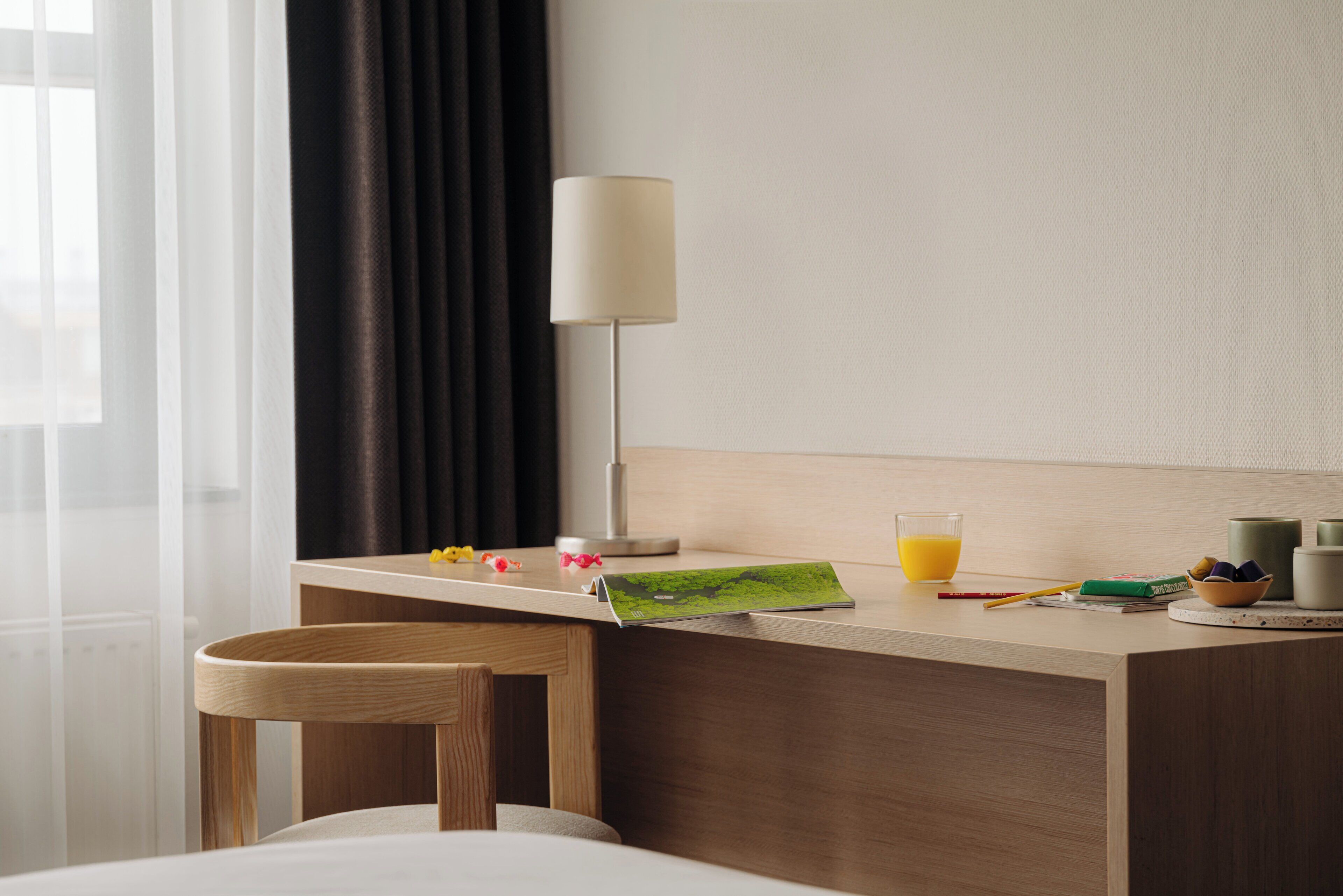 A hotel room with a wooden desk, a lamp, a glass of orange juice, and a small table with a bowl and a book.