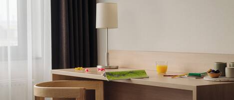 Quarto de hotel com uma escrivaninha de madeira, uma luminária, um copo de suco de laranja e uma mesinha com uma tigela e um livro.