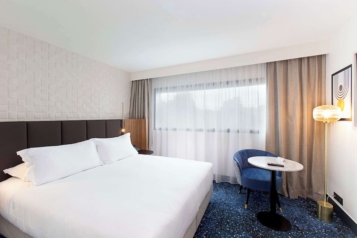 Une chambre d’hôtel avec un grand lit, une petite table ronde, une chaise et une grande fenêtre avec des rideaux.